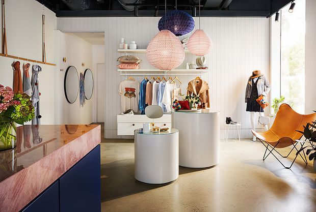 Bright, stylised retail clothing boutique with white vertical wall panelling, open shelving displaying garments and accessories, curved white display counters, a polished stone reception or product counter, colourful oversized pendant lights, a polished concrete floor, and a mid-century leather lounge chair by large windows. The exposed dark ceiling services add an industrial contrast to the playful, refined fit-out.