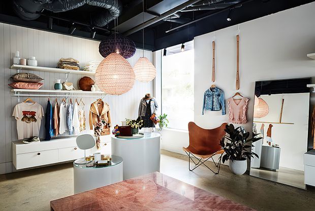 A bright, stylish retail clothing boutique with white vertical wall panelling, open shelving, hanging garments, display plinths, a large mirror, and a sculptural lounge chair. The space combines polished concrete flooring, a dark exposed services ceiling, woven pendant lights, and warm timber and leather accents.