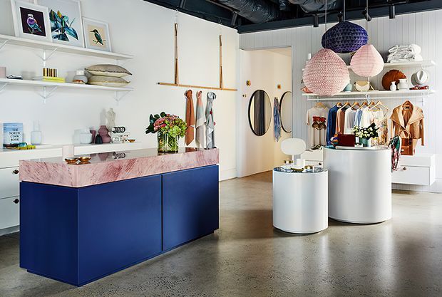 Bright contemporary retail showroom or boutique interior with polished concrete flooring, exposed dark ceiling services, white display shelving, clothing racks, colorful merchandise, and sculptural pendant lights. A bold navy-blue island counter with a pink veined stone top anchors the foreground, while white cylindrical display plinths and a curved white checkout or display counter occupy the centre and right side.