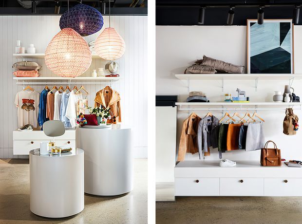 Bright contemporary retail clothing boutique interior with open white shelving, integrated hanging rails, drawers, folded garments, handbags, and display objects. The space features sculptural white cylindrical display plinths, polished concrete flooring, a ribbed white wall, exposed black ceiling services, and clustered woven pendant lights in warm and dark tones.