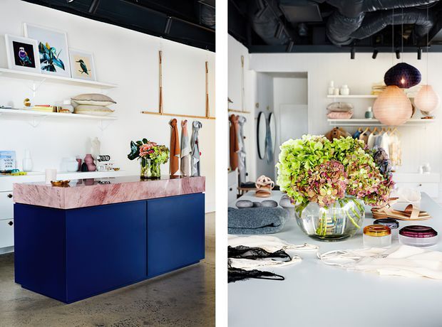 A bright, design-focused open-plan interior featuring a deep navy kitchen island with a pale pink marble benchtop, white open shelving, artwork, decorative objects, clothing rails, and a large floral arrangement. The space has an exposed dark ceiling with visible ductwork and layered pendant lights, combining workspace, display, and entertaining functions.