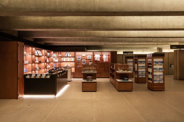 A warmly lit commercial retail interior with low exposed concrete beam ceilings, polished beige flooring, dark timber and reddish wood display cabinetry, illuminated counters, shelving, and freestanding product displays. The space has a refined museum or boutique showroom character with a deep, warm colour palette.