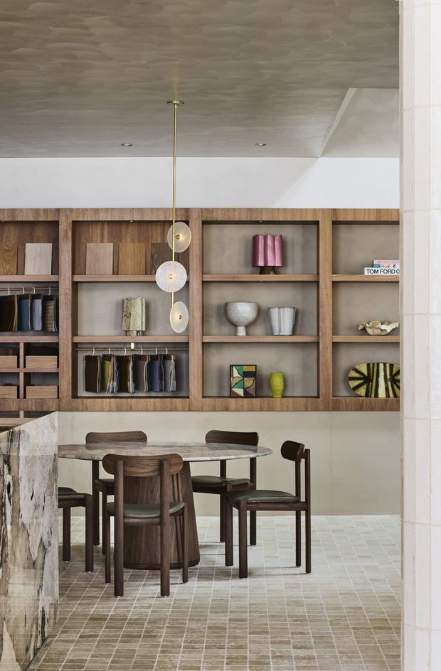 Refined dining area with a round stone-topped table and dark timber dining chairs, set against a full wall of warm timber display shelving and cabinetry. The shelving contains books, ceramics, artworks and decorative objects. A sculptural cluster pendant hangs above the table. Pale grid-pattern floor tiles, plaster walls and a textured ceiling create a warm, contemporary interior with an elegant material palette.