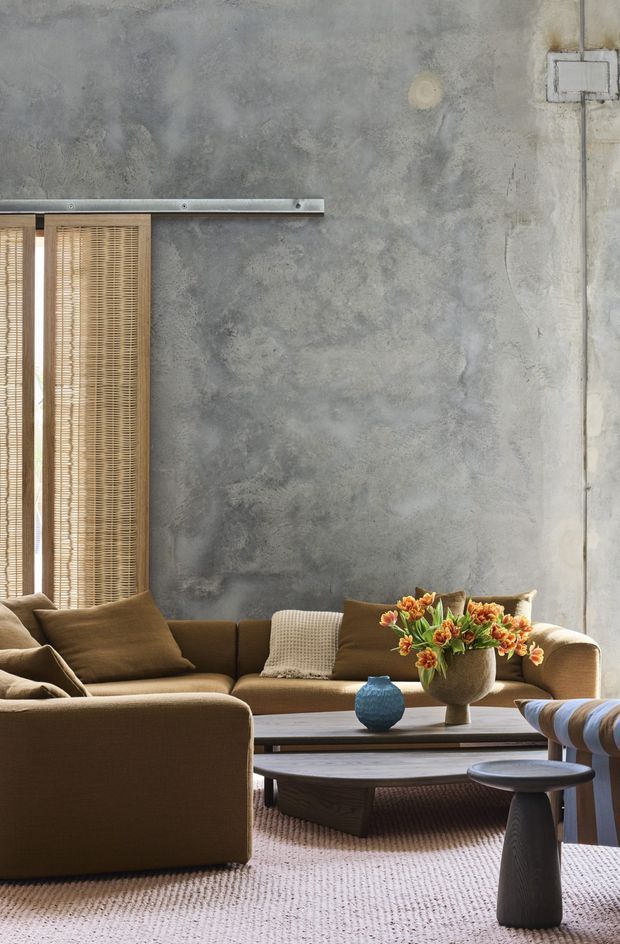 A warm, contemporary living room featuring a low-profile brown sectional sofa, textured neutral cushions, a large oval timber coffee table, and a dark sculptural side table. The room has a mottled grey concrete-look wall, a narrow window with timber slatted blinds, and soft natural light. A vase of orange flowers provides a small accent against the muted earthy palette.