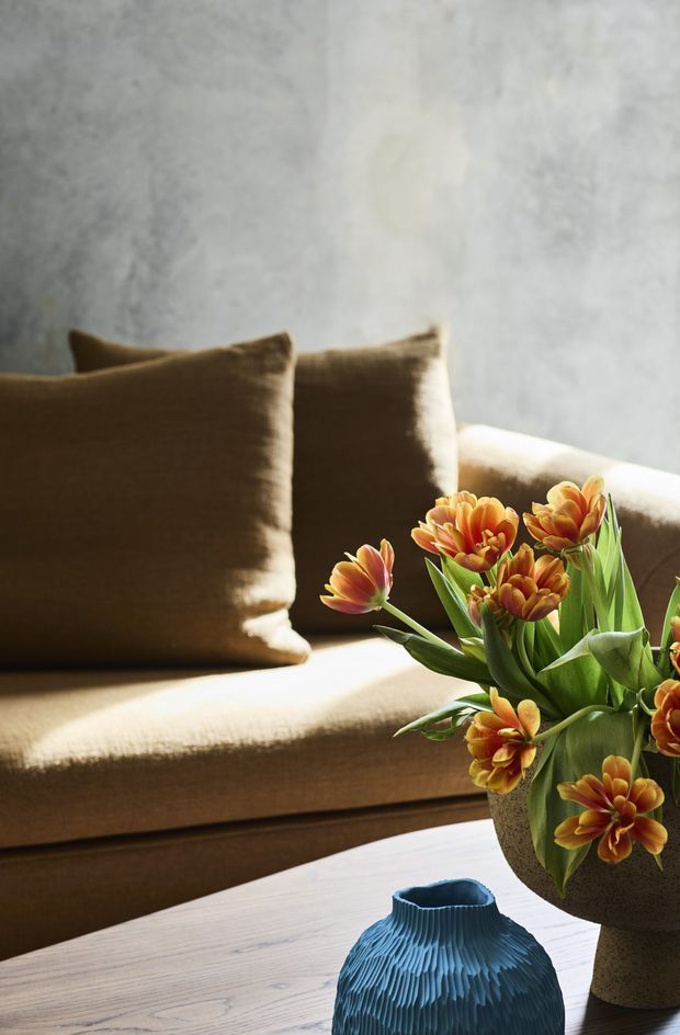 Close-up interior vignette of a warm brown upholstered sofa with matching cushions, a light timber coffee table, a textured blue decorative vase, and an arrangement of orange flowers in a neutral ceramic vessel. Soft natural light falls across a mottled grey feature wall and the seating, creating a calm, warm atmosphere.