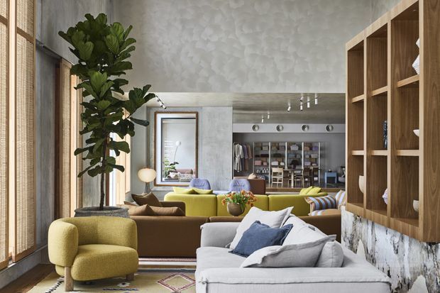 A richly layered open-plan living room with a low light-grey sectional sofa in the foreground, a mustard lounge chair, and a green sofa beyond. A tall indoor tree stands beside full-height windows with woven blinds. The room features a large timber open shelving unit with a marble-patterned base, textured pale walls, patterned rugs, decorative objects, and a glimpse of a dining or kitchen area through the opening at the rear. The style is eclectic and contemporary, with warm timber and earthy upholstery balanced by cool grey surfaces.