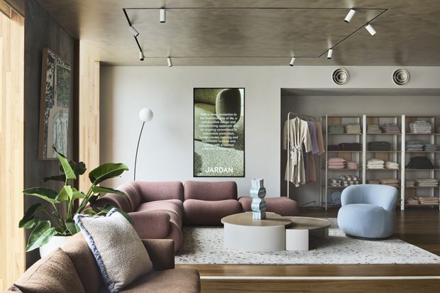 Contemporary open-plan living room with a sculptural mauve sectional sofa, pale blue lounge chair, round white coffee table, patterned rug, indoor plant, and framed artwork. A walk-in wardrobe or dressing area with open shelving and hanging clothes is visible behind the seating. The room has a textured concrete ceiling with recessed track lighting, warm timber wall and floor elements, and a calm neutral palette.