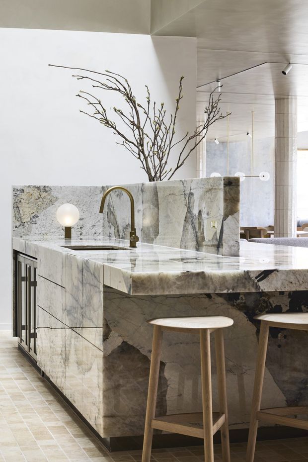 A refined open-plan kitchen centred on a large island clad in dramatic veined natural stone, with an integrated sink, brushed brass-toned gooseneck tap, pale cabinetry and timber bar stools. A low stone splashback partially screens the workspace, while a sculptural branch arrangement and spherical lamp add organic decorative elements. The kitchen opens to a contemporary living or dining area, with pale tiled flooring, a light-toned ceiling and slender recessed or track lighting.