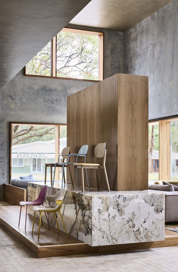 A dramatic contemporary interior with a raised timber platform and a large marble-clad architectural island or divider. The space features raw concrete walls and ceiling, expansive timber-framed windows, integrated timber joinery, bar-height seating, and sculptural chairs. A lower lounge area with a sofa is partially visible behind the stone-clad structure. The composition emphasizes layered levels, exposed materials, and abundant natural light.