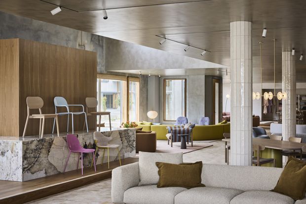 A spacious contemporary hospitality-style lounge and dining area with an open-plan layout. The room features a large curved light-upholstered sofa in the foreground, colourful lounge seating, a marble-clad bar or counter with timber cabinetry, polished concrete walls, timber ceiling panels, white tiled columns, and extensive windows. Track and pendant lighting add a refined modern character.