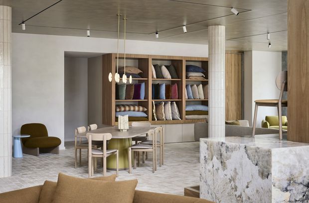 A refined open-plan interior combining a dining area, lounge seating, and a sculptural stone-clad kitchen or service counter. The space features pale textured walls, a grid-like tiled floor, a concrete-look ceiling with track lighting, fluted cylindrical columns, extensive warm timber built-in shelving filled with cushions, and a large heavily veined natural-stone island. Neutral upholstery and muted olive, blue, and rust accents create a calm, tactile atmosphere.