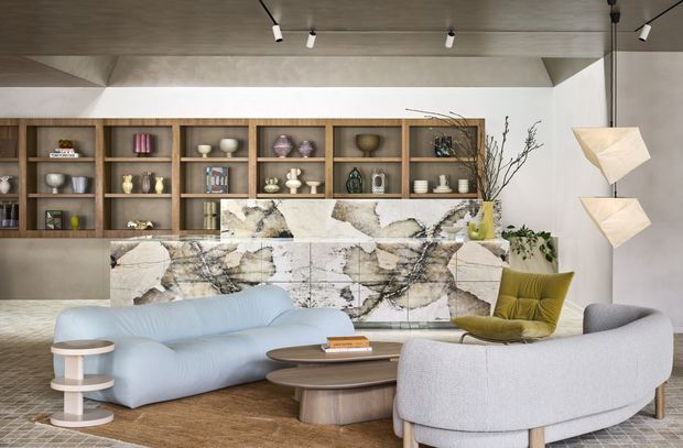 A sophisticated open-plan living room with a sculptural pale blue sofa, curved light-grey seating, layered oval coffee tables, and a mustard accent chair. The room features extensive built-in timber shelving displaying decorative objects, a dramatic veined stone-faced fireplace or media joinery wall, a polished concrete ceiling with track lighting, and sculptural pendant lamps. The palette is warm neutral with muted blue, olive, and natural timber accents.