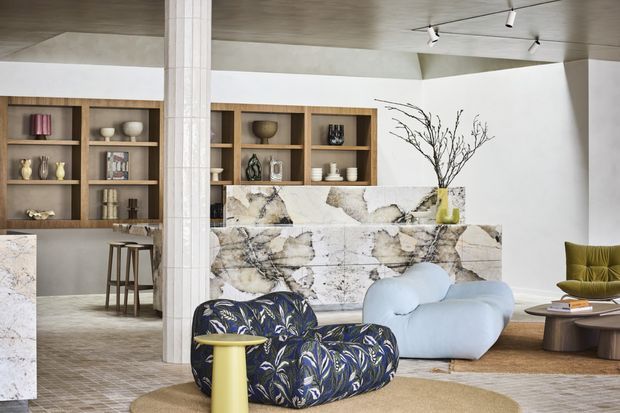 A sophisticated open-plan living and kitchen area with a sculptural marble-clad island, warm timber display shelving, and a contemporary seating arrangement. The room features a low concrete-toned ceiling, white walls, track lighting, a pale blue rounded sofa, a dark floral lounge chair, an olive green accent chair, and layered neutral rugs. A central structural column and built-in shelving contribute to the space's architectural character.