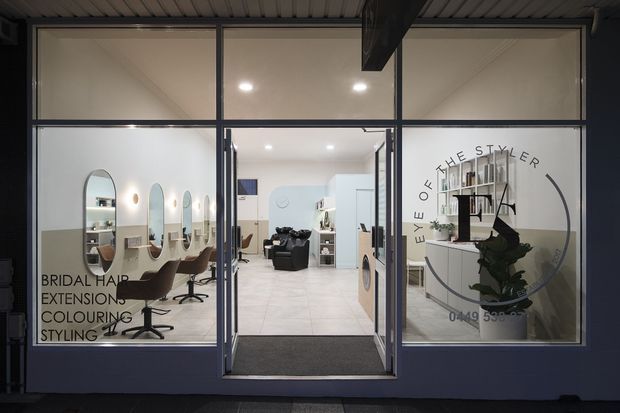 Front-facing view through a glazed storefront into a bright, contemporary hair salon. The salon contains styling stations with mirrors and chairs on the left, a shampoo basin and salon seating toward the rear, pale tiled flooring, white walls, and shelving with plants on the right. Black metal-framed glazing and a prominent salon logo occupy the facade.