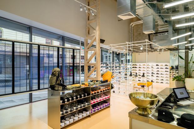 Bright contemporary retail showroom with exposed industrial services, polished concrete-style flooring, extensive glass frontage, and open shelving displaying footwear. A central island displays accessories, while a checkout counter and decorative planter occupy the foreground.