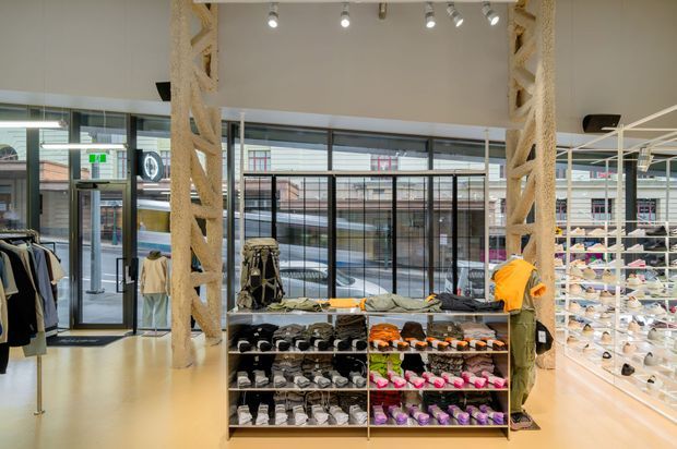 Bright contemporary retail footwear and apparel store with exposed structural columns, full-height glazed storefronts, track lighting, open shelving displays, clothing racks, and central shoe merchandise fixtures.