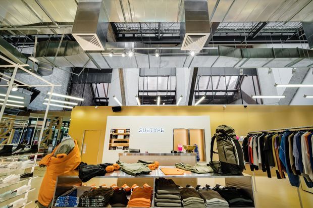 A brightly lit retail clothing store with exposed industrial ceiling services, large HVAC ducts, track lighting, clothing racks, display tables, shelving, and a central checkout or service counter. The space features a bold mustard-yellow partition wall and visible Subtype branding.