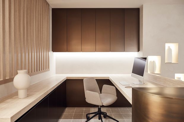 A refined built-in home office with a U-shaped dark timber desk and cabinetry, a pale stone-look work surface, integrated computer station, upholstered swivel chair, warm vertical timber wall battens, and softly illuminated display niches. The neutral, warm-toned interior has concealed LED lighting and a polished contemporary appearance.