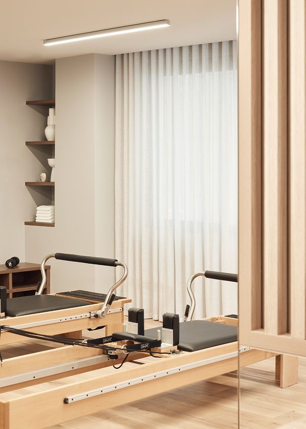 A bright, refined home Pilates studio with two wooden Pilates reformer machines in the foreground, a full-height mirrored wall on the right, sheer floor-to-ceiling curtains, pale timber flooring, built-in open shelving with neatly arranged towels and ceramics, and a slim linear ceiling light. The space has a calm, warm, minimalist feel with light natural materials and neutral tones.