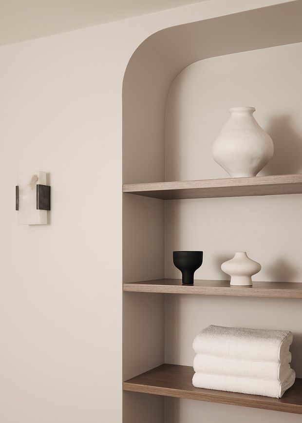 A close-up of a softly styled built-in arched shelving niche. The warm neutral plaster surround contains three timber-toned shelves displaying sculptural ceramic vessels, a black decorative object, and folded white towels. A minimalist wall sconce is mounted on the adjacent wall. The composition is calm, refined, and monochromatic with rounded architectural detailing.