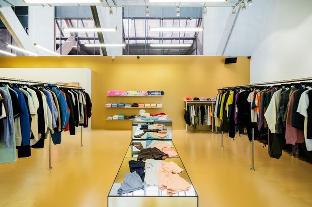 Bright contemporary clothing retail interior with a mustard-yellow feature wall, rows of garment racks, central glass display tables, open industrial-style ceiling services, and large overhead glazing.