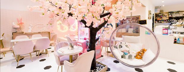 A whimsical pastel-themed commercial interior, likely a beauty salon or boutique, with blush-pink walls, pale pink upholstered seating, small round tables, a large artificial cherry-blossom tree, and a transparent hanging bubble chair filled with cushions and toys. Shelving and product displays line the rear wall, while the white floor features small dark geometric inlays. The space has a playful, highly decorative atmosphere with bright lighting.
