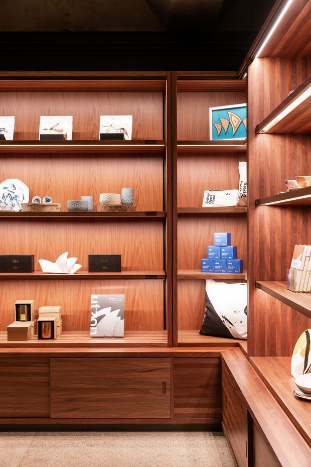 A warm, richly timber-lined retail or display space with extensive built-in open shelving, integrated cabinetry, and concealed linear lighting. The shelves hold books, ceramics, artwork, and small merchandise, while a low timber storage unit runs along the base. The space has a refined, gallery-like atmosphere with dark ceiling tones and a neutral carpeted floor.