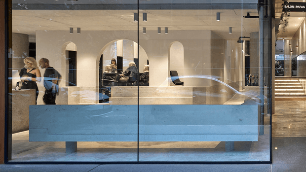 Street-level view through a full-height glazed storefront into a refined salon reception and styling space. A long pale blue reception counter dominates the foreground, with a warm neutral stone or plaster interior, arched openings, dark-framed glazing, pendant and recessed lighting, mirrors, salon stations, and a stepped illuminated corridor at the right. Reflections on the glass partially overlay the interior.