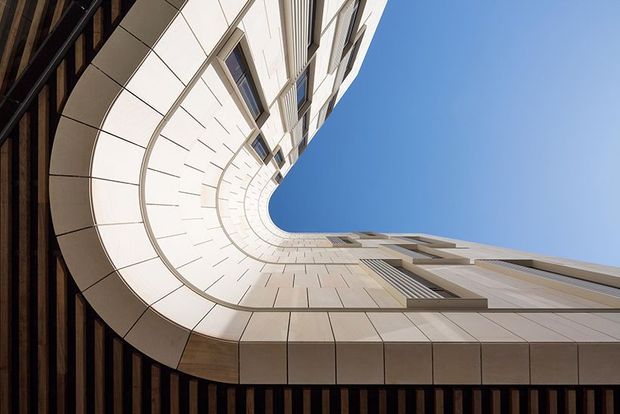 Upward-looking architectural view from within a narrow urban courtyard or building gap, showing a tall curved facade wrapping around an open blue sky. The facade combines pale rectangular cladding panels, projecting window bays, and dark vertical timber-like slats at the lower and left edges. Strong perspective, sweeping curves, and geometric repetition create a dramatic contemporary composition.