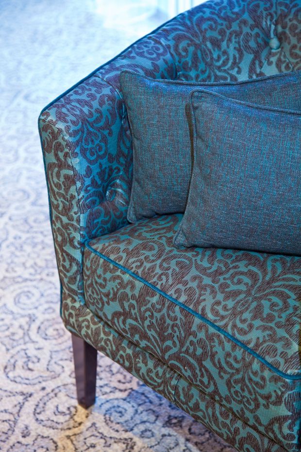 Close-up product-style photograph of a teal upholstered armchair with an ornate dark teal and brown patterned fabric, blue piping, matching textured cushions, and dark timber legs. The chair sits on a pale patterned rug; the image focuses on textile detail and furniture craftsmanship.