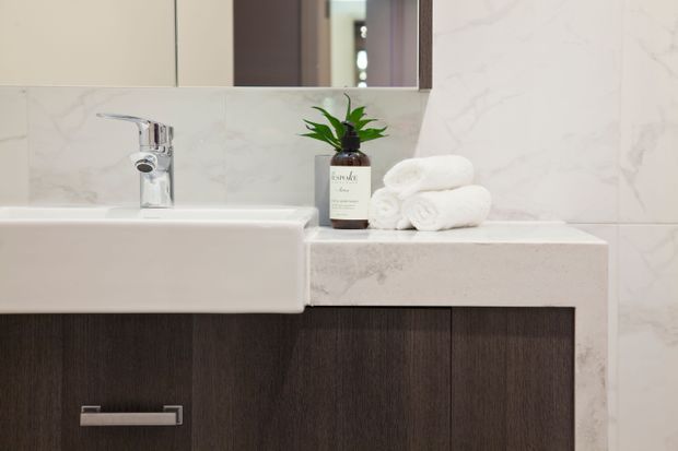 Close-up of a refined bathroom vanity featuring a white rectangular basin, chrome single-lever tap, pale marble-look wall and benchtop surfaces, and dark timber-grain cabinetry with a slim metal handle. A mirror, folded white towels, and a small plant complete the calm, clean composition.