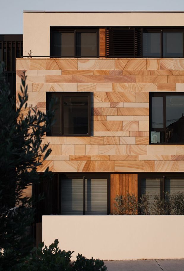Front-facing exterior view of a contemporary multi-storey residence. The facade combines pale rendered upper walls with extensive warm timber-toned patterned cladding arranged in rectangular panels, dark-framed windows, external louvres, and a timber entry door. A low rendered boundary wall and planted foreground frame the building.