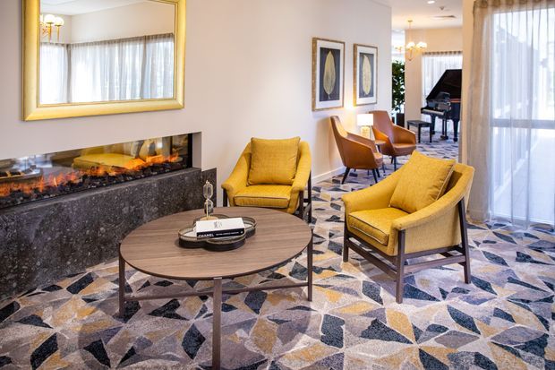 A refined living room or lounge with mustard-yellow upholstered armchairs arranged around a round dark wood coffee table. The room features a long built-in fireplace with a dark stone surround, patterned geometric carpet, sheer floor-to-ceiling curtains, framed artwork, a large gold-framed mirror, and a grand piano visible in the adjoining area. The palette is warm and elegant with mid-century-inspired furnishings and polished decorative detailing.