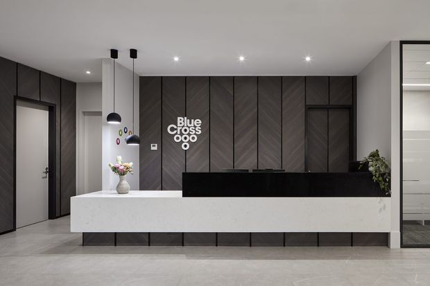 A contemporary commercial reception area with a long white stone reception desk, a black raised counter, and dark vertically panelled walls featuring the Blue Cross logo. The space has a polished light-grey floor, recessed ceiling lights, black pendant lights, integrated dark cabinetry or wall panels, and glazed office or meeting-room doors at the sides.