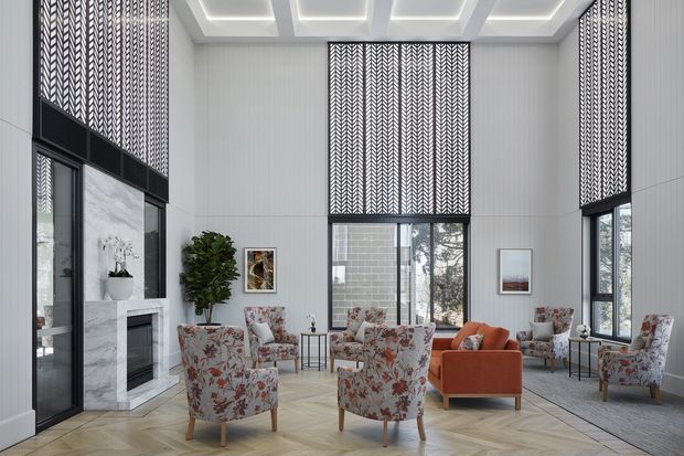 A bright, double-height sitting room with large black-framed windows and patterned roller blinds. The room features a white marble fireplace, pale wall panelling, a coffered ceiling with recessed lighting, herringbone timber flooring, floral upholstered armchairs, and a vivid orange sofa arranged around small occasional tables.