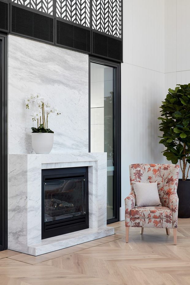 A refined living room corner featuring a white and grey-veined marble fireplace surround with a black inset firebox, tall dark-framed glazing, decorative geometric ventilation panels, and pale timber herringbone flooring. A floral upholstered armchair, potted orchid, and large indoor tree add softness and colour. The space has a polished, contemporary feel with strong black-and-white contrast and warm wood flooring.