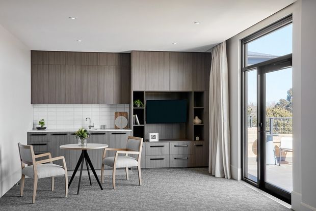 A bright, compact open-plan living and dining area with a contemporary built-in timber entertainment and storage wall, a small round dining table with upholstered chairs, a compact kitchenette with white tiled splashback, and full-height black-framed glazing opening to an outdoor terrace. Neutral finishes, recessed ceiling lights, and sheer curtains create a refined, understated interior.
