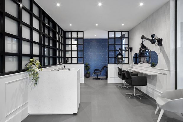 A bright, modern hair salon interior with a white reception counter in the foreground, black-framed glazed partitions and doors, grey flooring, a blue geometric feature wall, and a long styling counter with black salon chairs and wall-mounted mirrors. White paneled walls and sculptural pendant-style salon lighting create a refined contemporary commercial fit-out.