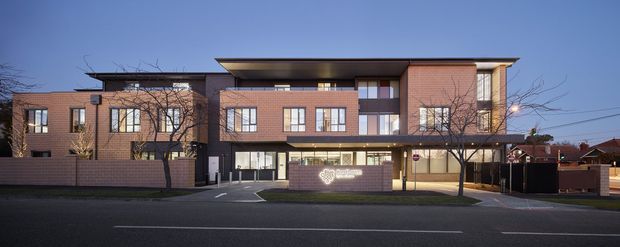 Street-facing exterior photograph of a multi-storey contemporary brick building at dusk. The facade combines warm red brick, dark charcoal cladding, broad horizontal roof overhangs, and extensive rectangular glazing. A low brick boundary wall and illuminated entry signage define the frontage, with a covered ground-level entrance and side driveway visible.