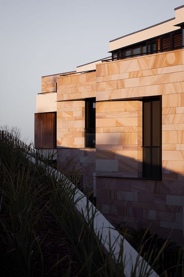 Exterior architectural photograph of a contemporary multi-level residence clad in warm, horizontally laid timber-toned panels. The facade features strong geometric volumes, dark recessed window and door frames, upper-level louvres or screens, and flat roof forms. Dense ornamental grasses occupy the foreground, while low-angle sunlight creates pronounced shadows across the facade.