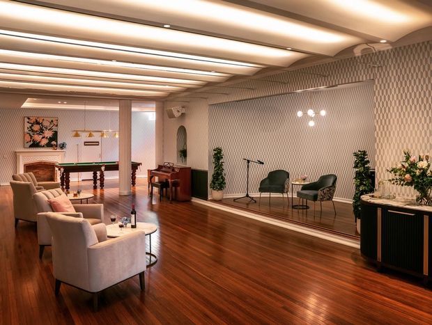 A refined mid-century-inspired lounge and entertaining space with polished dark timber flooring, upholstered armchairs, a pool table, piano, decorative fireplace, and a raised sitting area. The room features a patterned textured wall finish, mirrored or glossy detailing, a long built-in ceiling with recessed linear lighting, and warm ambient illumination. A console with floral arrangements occupies the foreground.