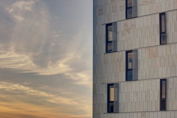 Close-up exterior architectural view of a multi-storey building facade clad in pale rectangular stone or panel modules, with narrow dark-framed windows and recessed vertical openings. The facade fills the right side of the image while a warm, cloud-filled sunset sky occupies the left, creating a minimalist contemporary composition.