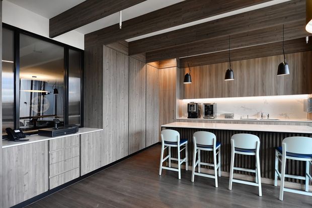 A contemporary kitchen and entertaining space dominated by dark timber cabinetry and wall panelling. A long island with a pale stone benchtop and integrated sink is paired with four white bar stools, while black pendant lights hang above. Floor-to-ceiling timber joinery, dark flooring, black-framed glazed doors, and warm concealed lighting create a refined, high-contrast interior.