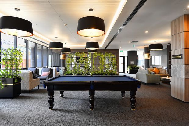 A contemporary communal lounge or recreation area featuring a full-size black billiards table in the foreground, surrounded by sofas and armchairs. Large glazed windows line the left side, while a planted green wall and indoor planters add natural elements. The space has a carpeted floor, suspended black drum pendant lights, recessed lighting, timber and stone wall joinery, and a long linear ceiling detail. The setting resembles a refined hospitality, workplace, or apartment amenity space.