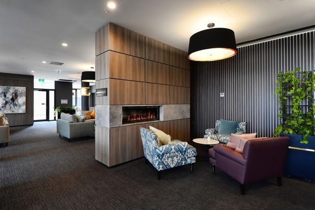 A contemporary commercial lounge or reception area with a built-in linear fireplace set into a timber-panelled feature wall. The space includes patterned upholstered armchairs, a deep purple lounge chair, a small round table, dark carpet, pendant lighting, a vertically ribbed dark feature wall, and integrated greenery. A lobby or entry zone is visible in the background.