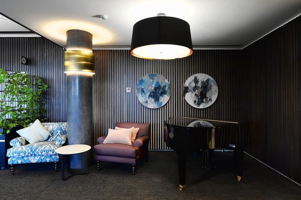 A dark, dramatic lounge or reception area with black vertical timber wall panelling, charcoal carpet, sculptural pendant lighting, a cylindrical fireplace or column with glowing horizontal bands, patterned blue seating, a mauve armchair, and a black grand piano. Abstract circular artwork and indoor greenery add decorative elements.