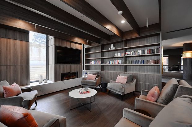 A sophisticated living room with dark timber built-in cabinetry, full-height bookshelves, a wall-mounted television, and a linear fireplace set into a pale stone surround. Several upholstered lounge chairs and sofas with blush cushions are arranged around a round coffee table. Dark exposed ceiling beams and integrated lighting create a warm, refined atmosphere, while glazing at the rear provides natural light and a view toward an adjoining interior space.