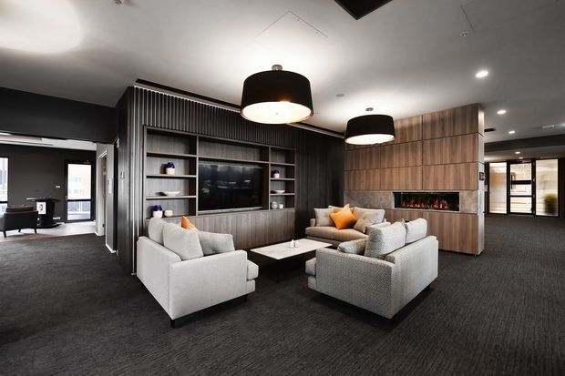 A sophisticated contemporary lounge area with two pale upholstered sofas and a pair of armchairs arranged around a low rectangular coffee table. The room features dark charcoal carpet and walls, a full-height dark timber entertainment and shelving unit, and a matching timber fireplace surround with a linear fire. Large black drum pendant lights hang over the seating area, while glazed doors and windows provide views into adjoining spaces. The overall design is polished, warm, and high contrast.
