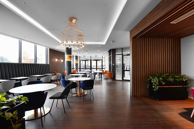 A contemporary commercial café or hospitality interior with an open dining area, round tables, upholstered chairs, a long dark banquette, and a dramatic cluster of brass pendant lights. The space features dark timber flooring, a ribbed white ceiling with concealed lighting, a substantial vertical timber slat feature wall and ceiling detail, black-framed glazing and doors, and integrated planter boxes. The bright, polished interior has a refined modern atmosphere.