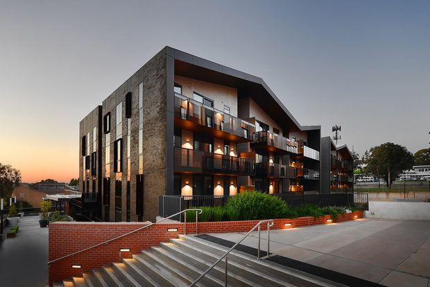 Dusk exterior view of a contemporary multi-storey apartment or townhouse building. The facade combines dark grey metal framing and cladding with exposed reddish-brown brick, extensive balconies with glass and dark metal balustrades, and warm illuminated interiors. Broad concrete paving, landscaped raised planting beds, brick retaining walls, stairs, and stainless-steel handrails occupy the foreground.